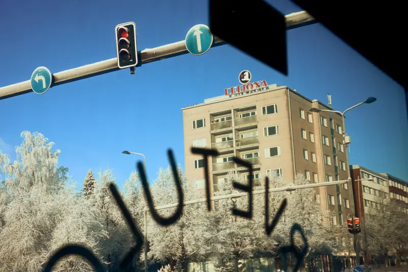 A winter street scene viewed through a frosty bus window. Condensation or frost on the glass spells a word in reverse. Outside, under a clear blue sky, frost-covered trees line the street next to a traffic light. In the background stands an apartment building topped with a red neon sign reading 'LEIJONA' next to a lion logo.