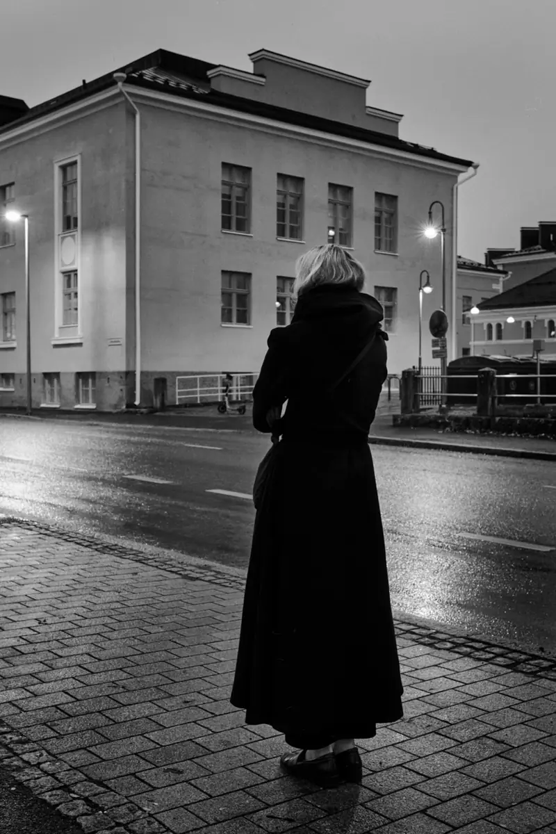 A black and white street photograph taken at dusk. A person in a long dark coat stands with their back to the camera, looking across a wet, streetlight-illuminated road towards a large, multi-story building with a classic facade.