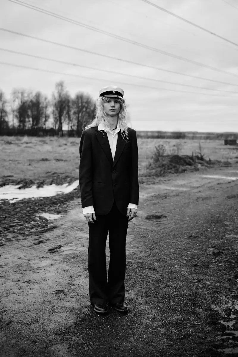 A full-body black and white portrait of a recent graduate in a traditional Finnish graduation cap and a dark suit. They stand on a muddy dirt path in an open landscape with bare trees and overhead power lines under a cloudy sky.