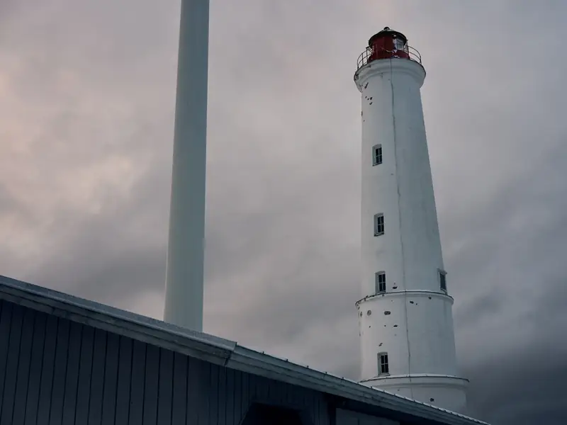 A photograph of the historic Marjaniemi lighthouse in Hailuoto, Finland. The tall, white brick lighthouse with its red lantern room stands against an overcast, grey sky. A sleek, modern white wind turbine tower is visible to the left, and the edge of a dark wooden building frames the bottom.