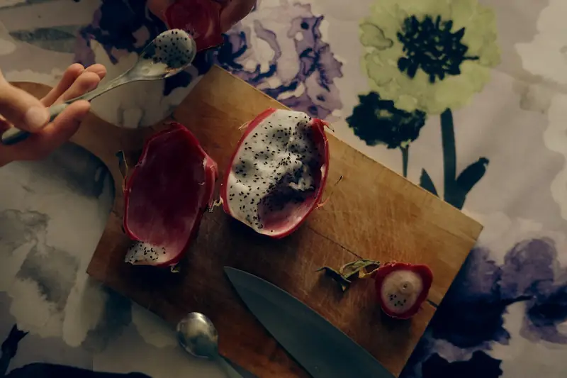 A top-down photograph of a wooden cutting board on a floral tablecloth. On the board is a halved dragon fruit (pitaya) with vibrant pink skin and white flesh speckled with black seeds. A hand is holding a spoon, scooping out a piece of the fruit, while a kitchen knife rests nearby.