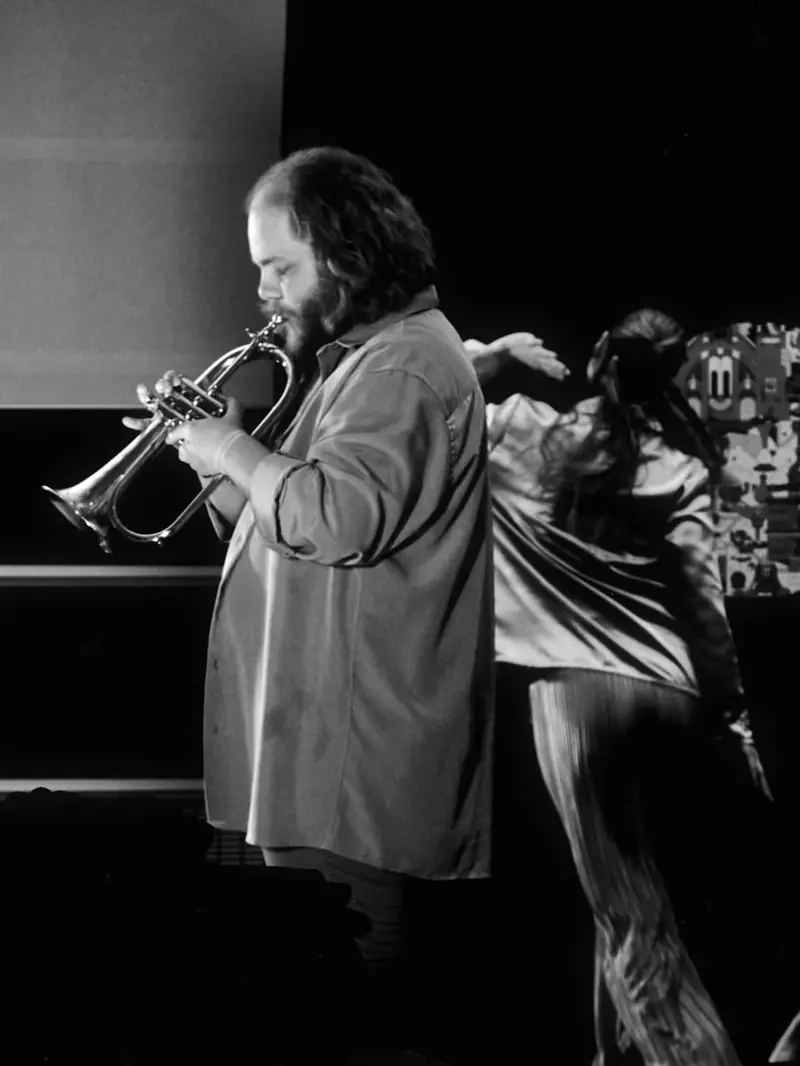 A black and white photograph of a live musical performance. In the foreground, a bearded man plays a flugelhorn with intense focus. Behind him, slightly out of focus, a woman dances expressively in shiny clothing against a dark background.