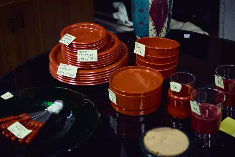 A close-up photograph of vintage red and orange plastic tableware for sale at a flea market. Stacks of plates and bowls, along with glasses and spoons, are arranged on a dark, glossy table. Small handwritten price tags in Finnish are taped to the items.