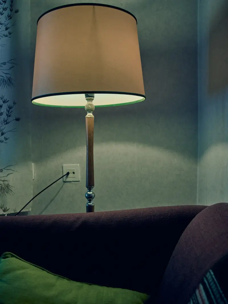 A lit floor lamp with a beige shade and wooden stand against a light blue-grey wall. The edge of a purple sofa with a green pillow is visible in the foreground.