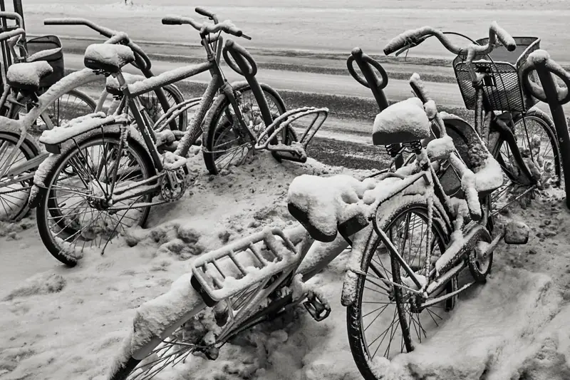 A black and white photograph showing a close-up of several bicycles parked haphazardly together in a deep snowbank. The bikes, including their seats, handlebars, and baskets, are heavily covered in thick, fresh snow. One bicycle in the foreground is partially tipped over.