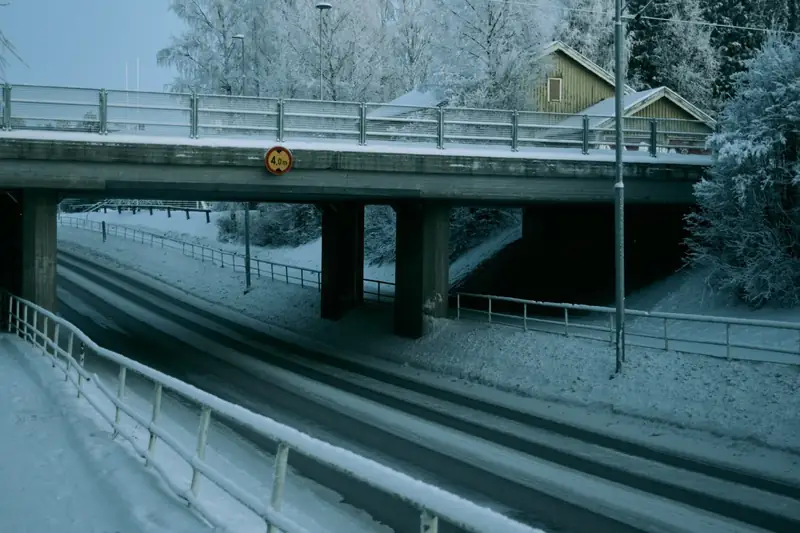 A cold winter photograph of a snowy road passing underneath a concrete overpass in Ylivieska, Finland. The surrounding trees and ground are covered in thick frost, and a yellow wooden house is visible in the background above the bridge.