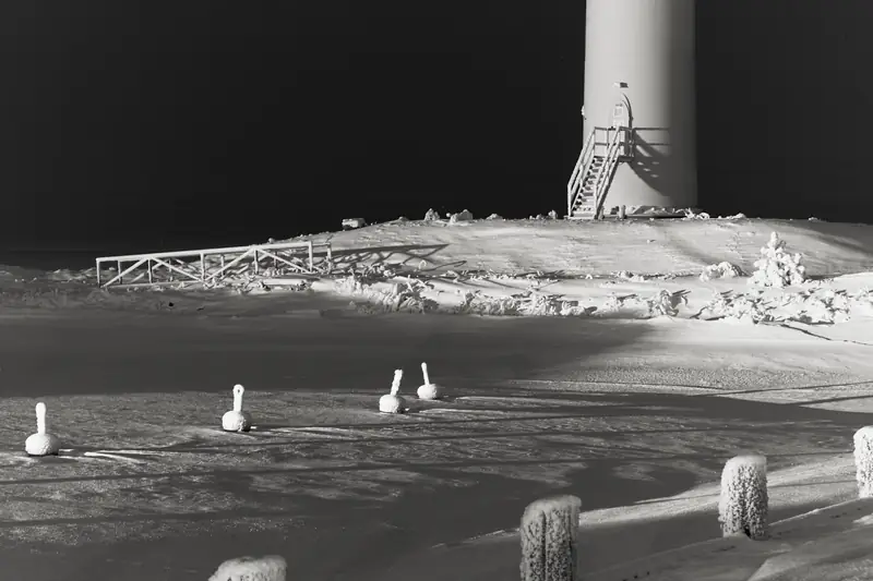 A high-contrast black and white winter landscape at night. In the foreground, small snow-capped buoys protrude from a frozen, flat surface. In the background, the base of a white lighthouse with a wooden staircase stands on a snow-covered shore against a pitch-black sky.