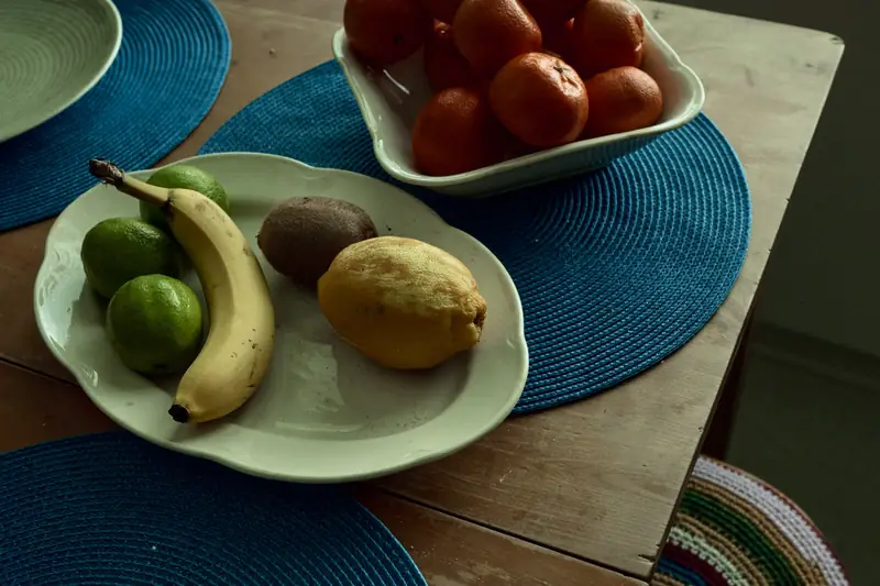 A still life photograph of fresh fruits on a wooden dining table. In the foreground, a white ceramic plate holds a yellow banana, limes, a kiwi, and a lemon. In the background, a white bowl is filled with vibrant oranges. The plates sit on bright blue circular woven placemats, captured in soft natural indoor light.