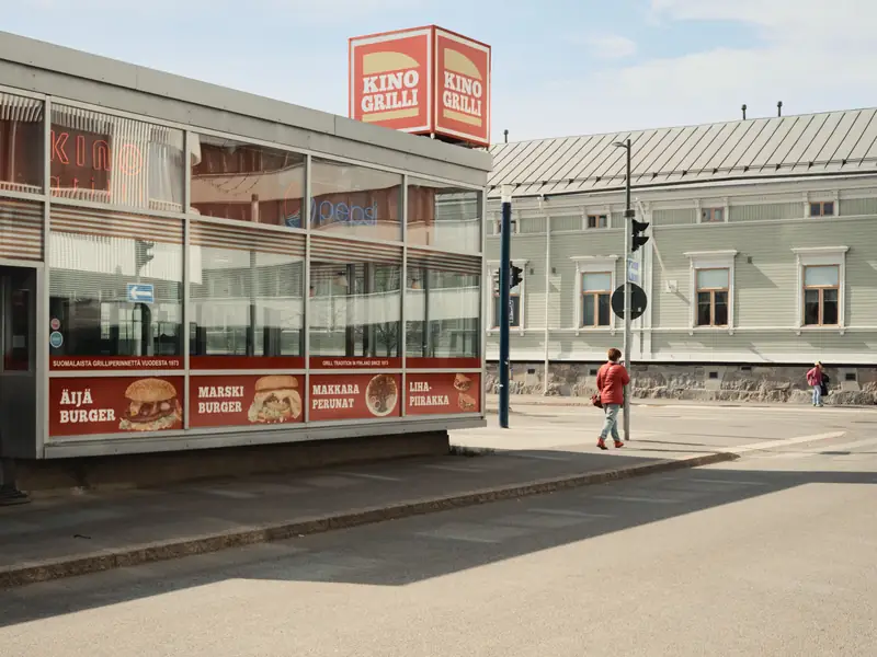 A sunlit street scene featuring a classic Finnish fast-food kiosk named 'KINO GRILLI' with its distinctive red and yellow sign. A person wearing a red jacket walks on the sidewalk past the kiosk's glass windows, which display advertisements for various burgers. A traditional light green wooden building stands in the background.