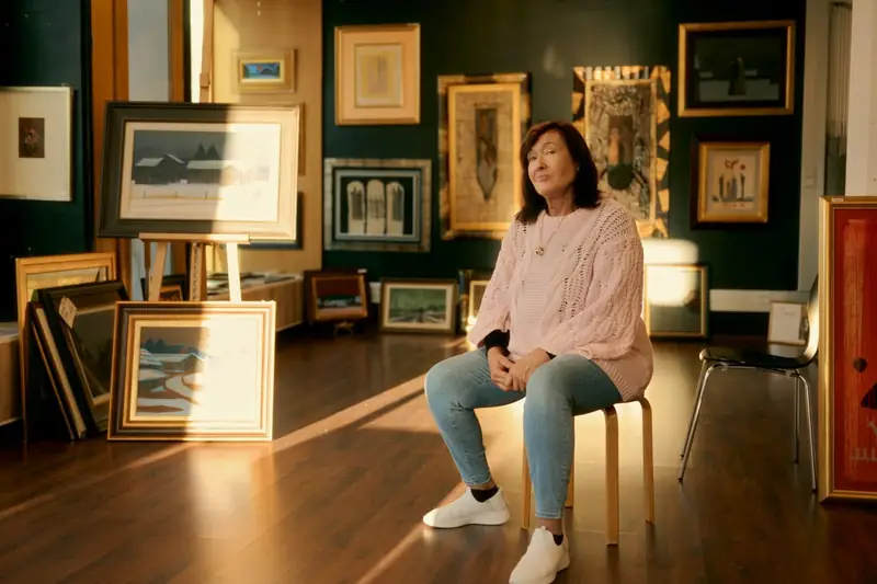 Leila Palmu, the widow of Finnish artist Juhani Palmu, sitting on a wooden stool inside the Torikatu gallery in Oulu, 2025. Warm evening sunlight crosses the wooden floor and illuminates her pink sweater. Framed paintings hang on the dark green walls behind her.