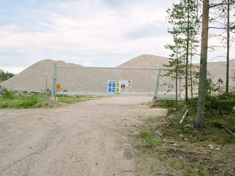 A wide landscape photograph showing the entrance to an open sand and gravel quarry in Murto, Tyrnävä. A closed light green metal security gate with safety warning signs blocks a dirt road in the foreground. Behind the gate, massive smooth piles of extracted gravel rise against an overcast sky.