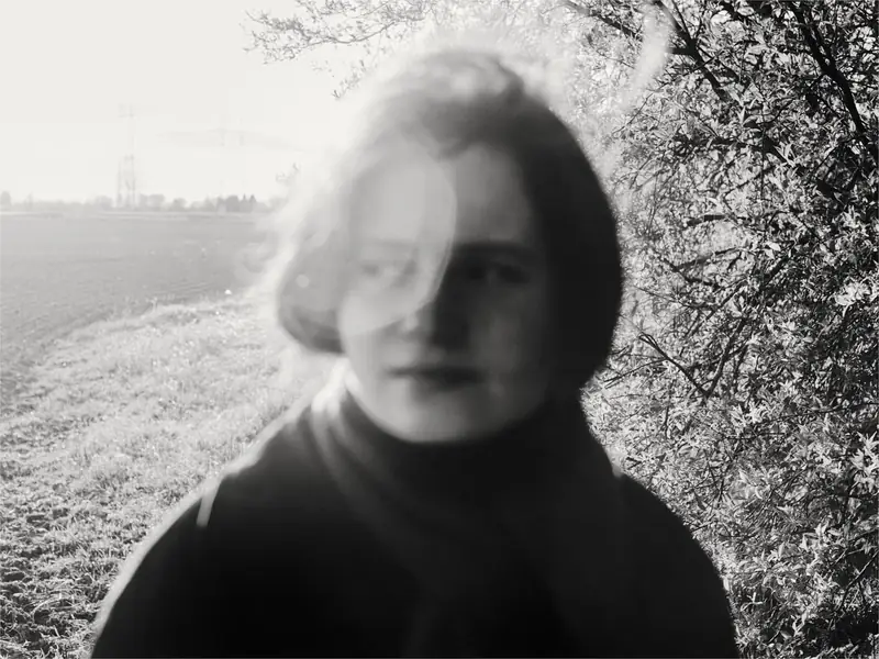 A black and white portrait of a young person outdoors. A light leak or lens flare partially covers their face. They look to the side, standing between an open field and a bare bush.
