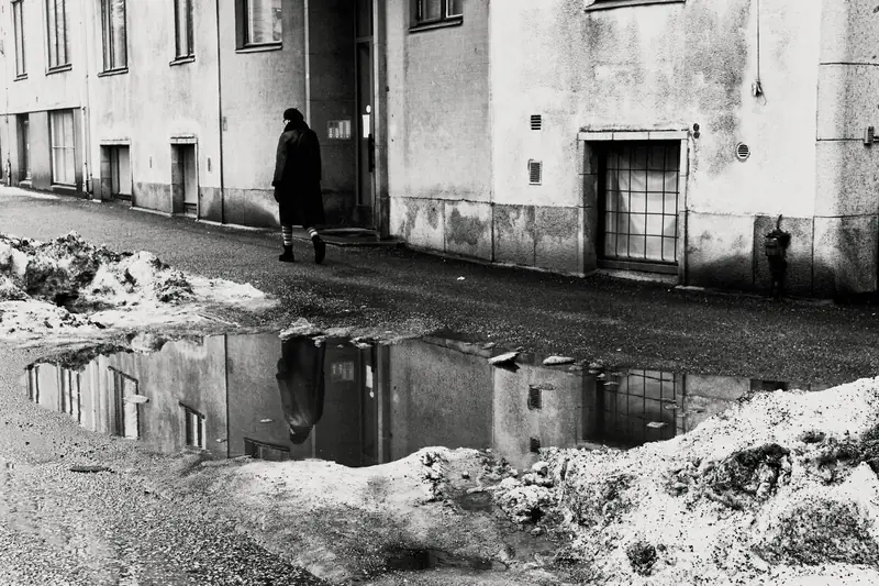 A high-contrast black and white street photograph of a person in a dark winter coat walking down a street in Helsinki. In the foreground, a large puddle on the asphalt perfectly reflects the person and the textured facade of the residential building behind them, framed by melting snow piles.