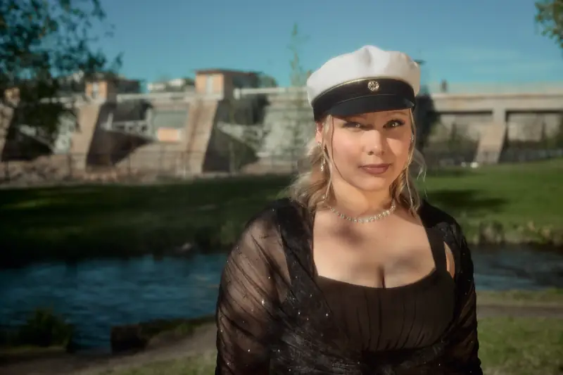 Close-up portrait of a person wearing a traditional Finnish graduation cap (Ylioppilaslakki) and a black outfit, standing outdoors. In the background, the Oulu dam is visible in soft focus, situated near the subject