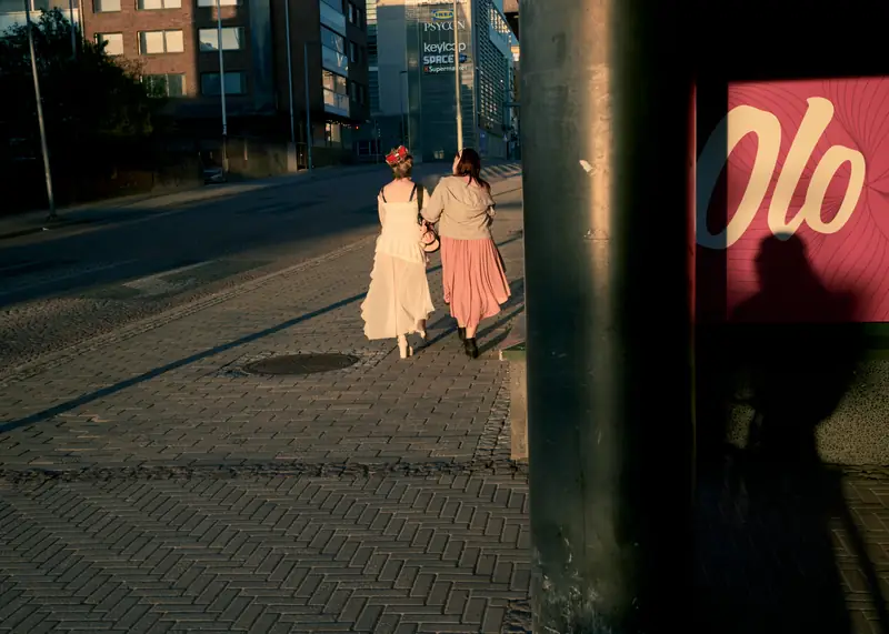 A golden hour street photograph taken from behind two women walking arm-in-arm down a cobblestone sidewalk in Oulu, Finland.
