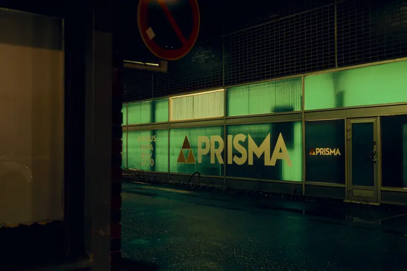Prisma supermarket at night. The glass facade glows green from inside, and the light reflects off the wet street in front of the building.