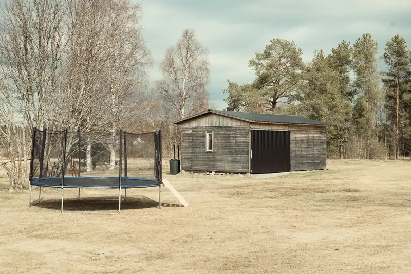 A landscape photograph of the same rustic wooden outbuilding, now in early spring or autumn. The snow has completely melted, revealing a field of dry, yellow-brown grass. A large black trampoline stands prominently in the foreground on the left. The surrounding trees are still bare, set against a bright, partly cloudy sky.