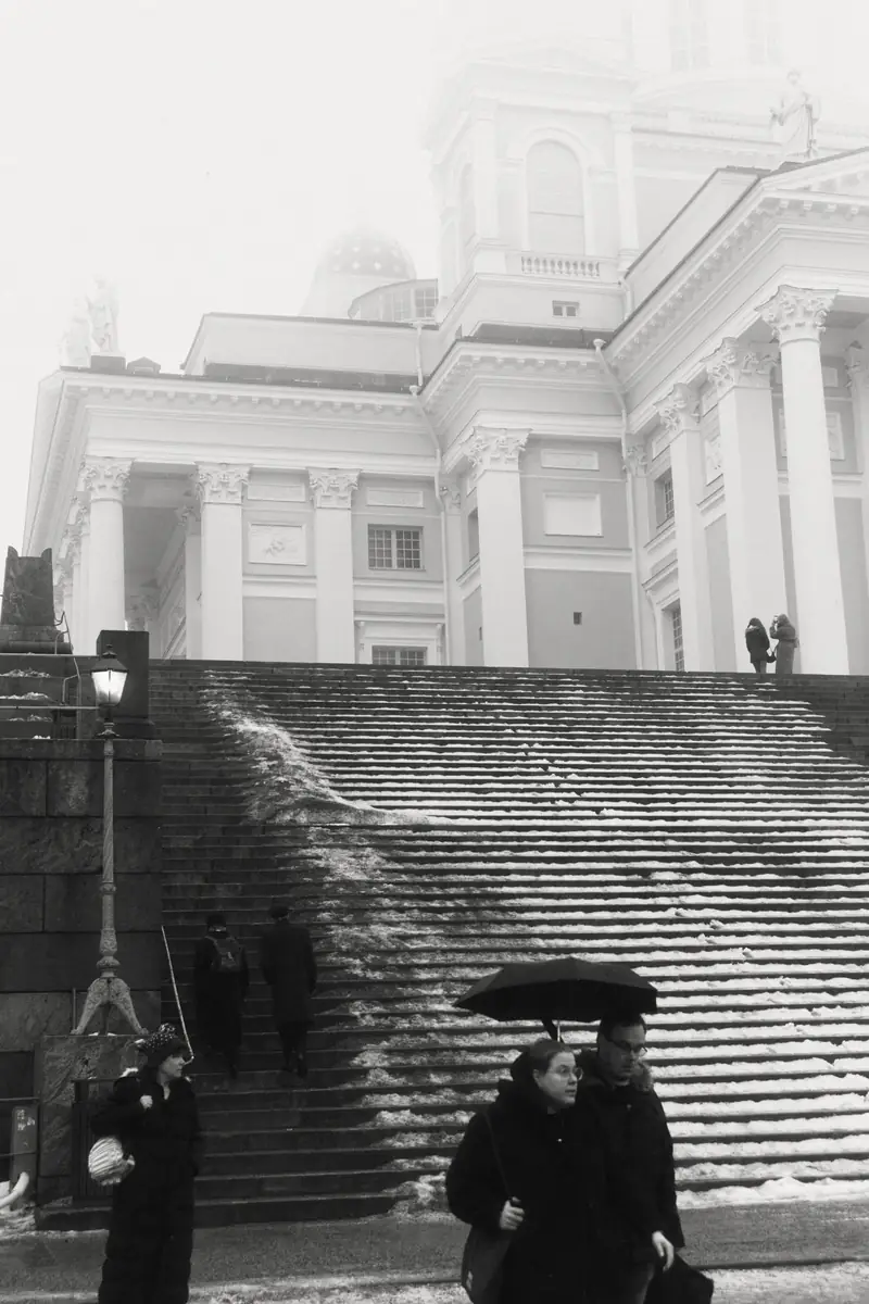 A black and white photograph of the Helsinki Cathedral partially obscured by thick fog on a winter day. The grand white columns contrast with the dark winter clothing of several people walking on the wide, snow-covered stone stairs in the foreground, including a couple sharing an umbrella.