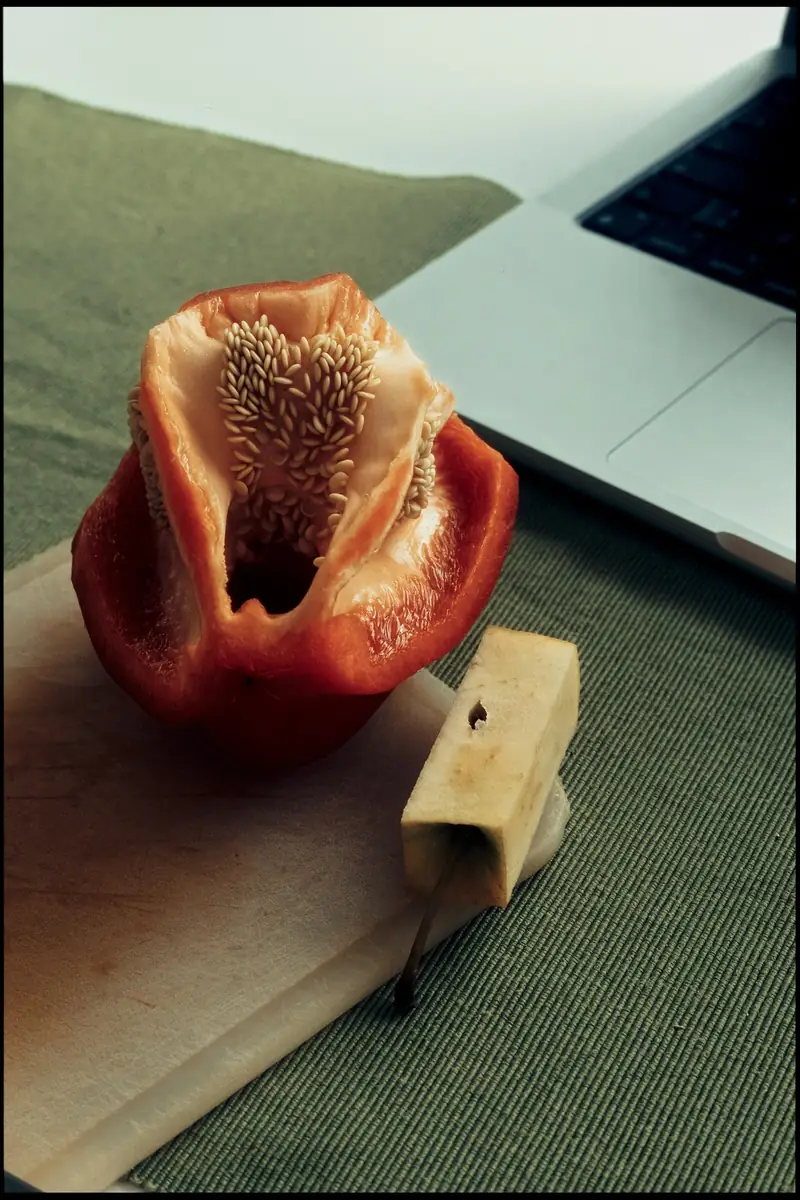 A moody still life photograph of an opened red bell pepper revealing its seeds, resting on a white cutting board over a green textured placemat. Next to it is a square-cut apple core with its stem. A silver laptop is partially visible in the background.