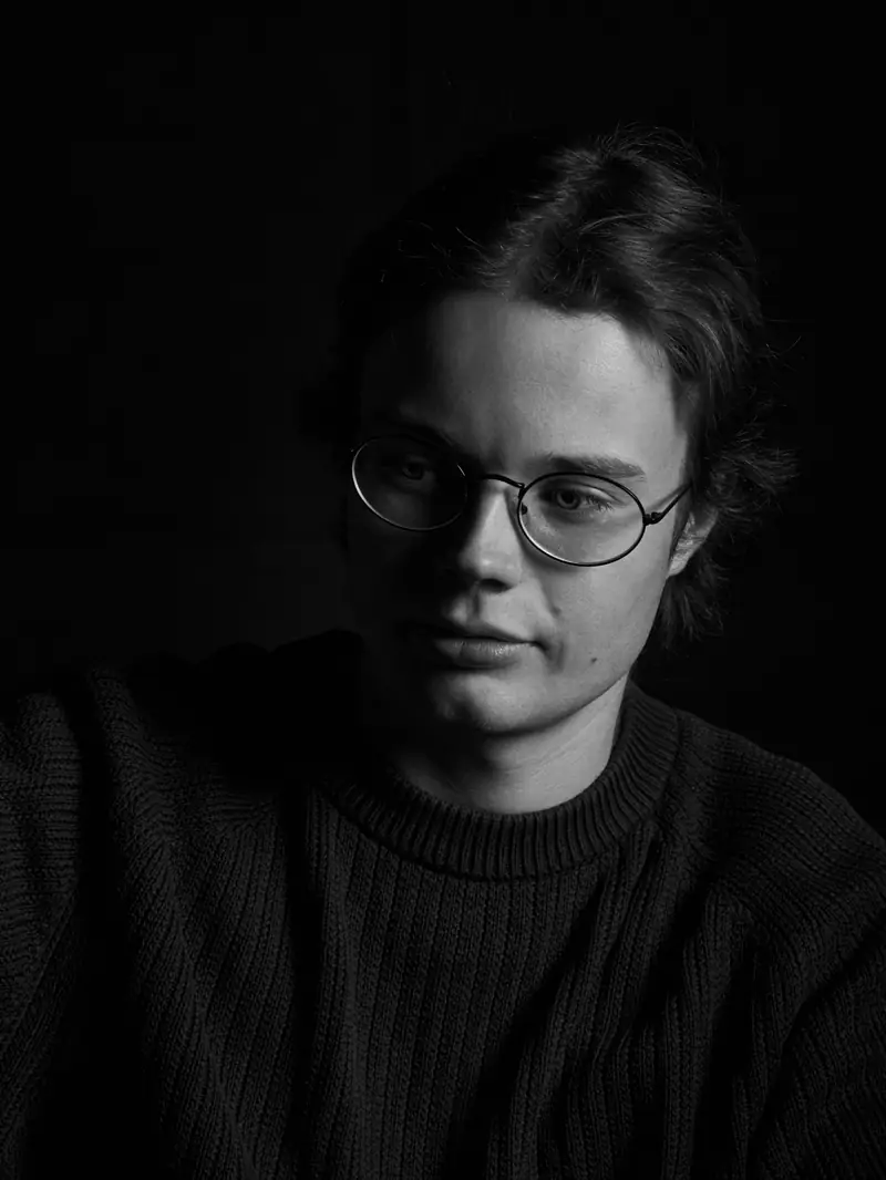 Black and white studio portrait of a young man with glasses wearing a dark knitted sweater. He looks to his right, lit by strong side lighting against a black background.