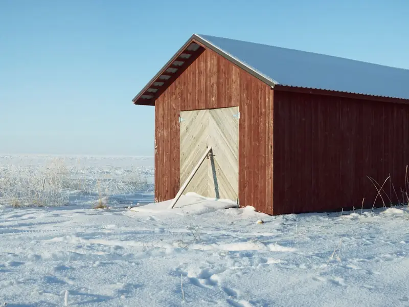 A small, bright red wooden shed with a snow-covered roof and light wooden double doors. Flat, snow-covered ground around it and a clear pale blue winter sky behind.