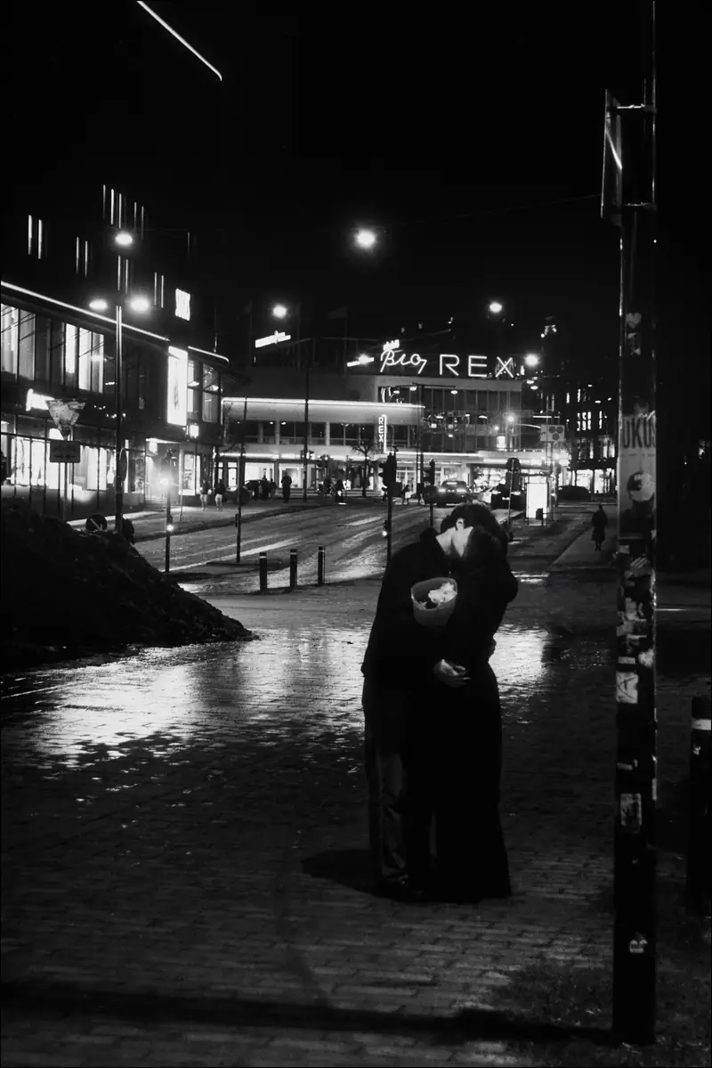 A couple kissing on a wet city street at night. One holds a bouquet of flowers. Neon signs including one for 'Bio REX' glow in the background and reflect off the rain-slicked pavement.